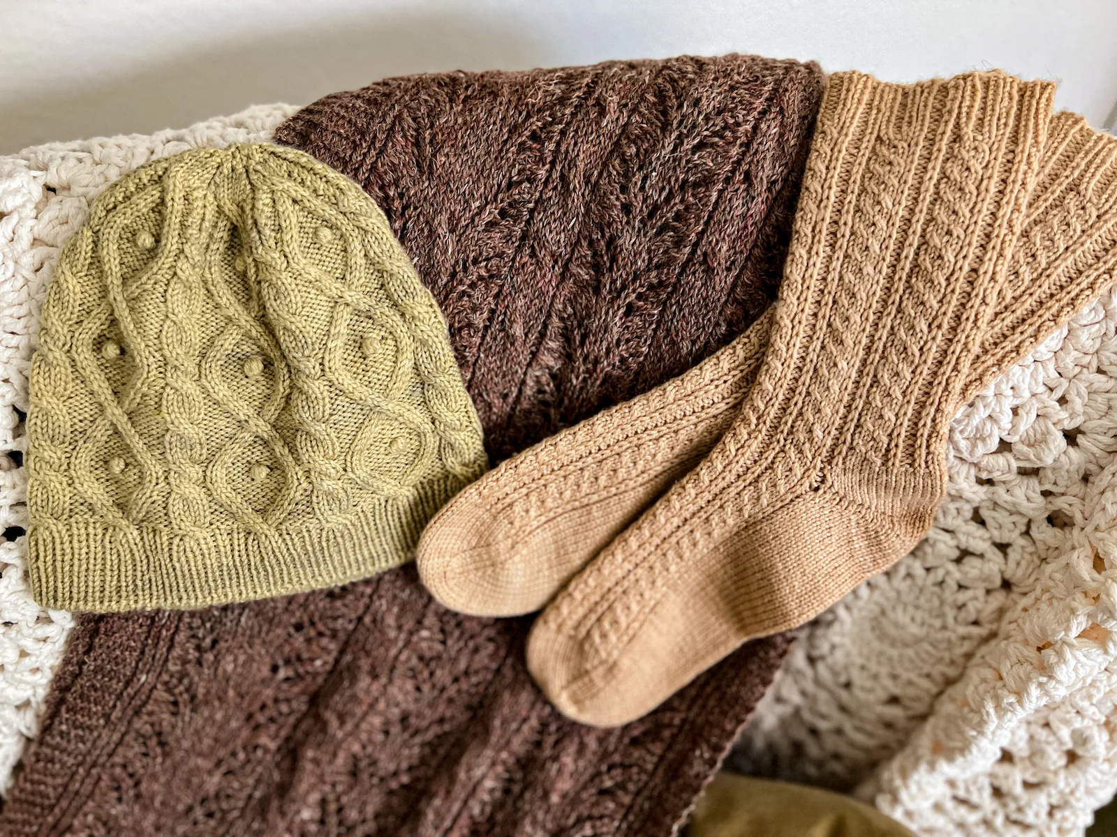 A light green knit hat, a brown knit wrap, and a pair of peach knit socks are all displayed on the back of a chair. Each of the knit items has lots of different styles of cables.