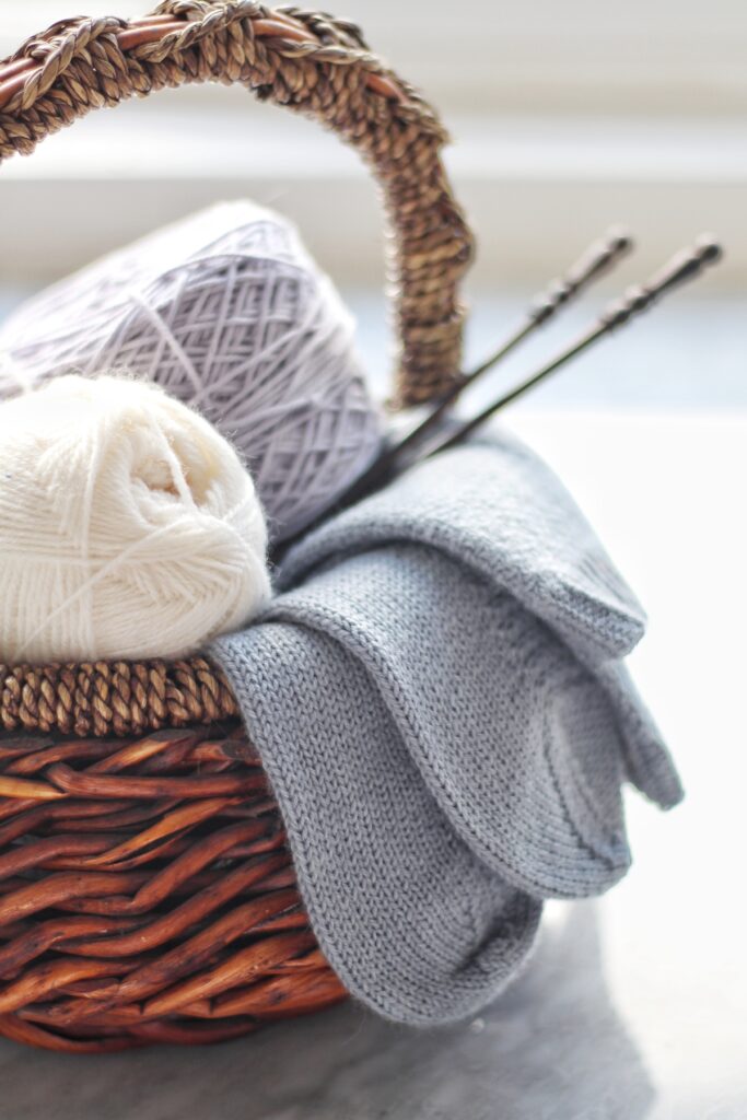 A brown basket full of white and lavender yarn, a pair of light blue knit socks hanging over the side, and a pair of dark brown straight knitting needles sticking out to the right.