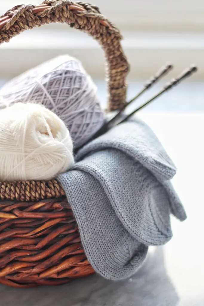 A brown basket full of white and lavender yarn, a pair of light blue knit socks hanging over the side, and a pair of dark brown straight knitting needles sticking out to the right.