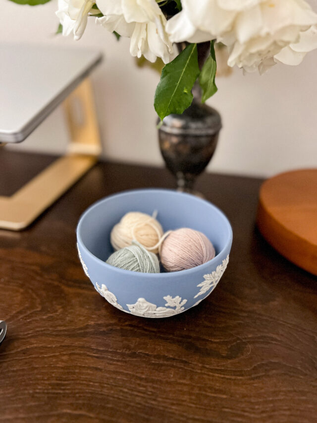 Three small balls of yarn sit inside a light blue jasperware bowl. The bowl sits on a dark wood desk surrounded by a laptop and a vase of flowers.