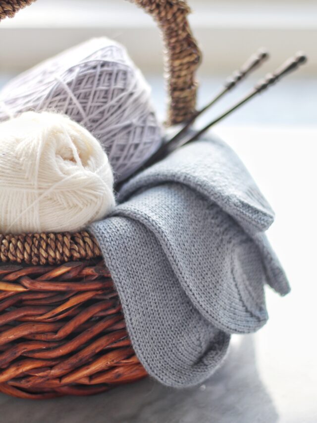 A brown basket full of white and lavender yarn, a pair of light blue knit socks hanging over the side, and a pair of dark brown straight knitting needles sticking out to the right.