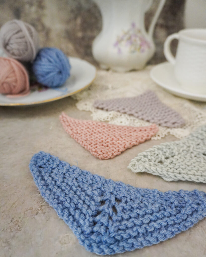 A close-up on a light blue triangular mini shawl. This shows a variation on the traditional symmetrical triangle with center increases, where additional increases are worked at the beginning and end of each wrong side row to crease a shawl that grows outward faster and has a shallower center point.