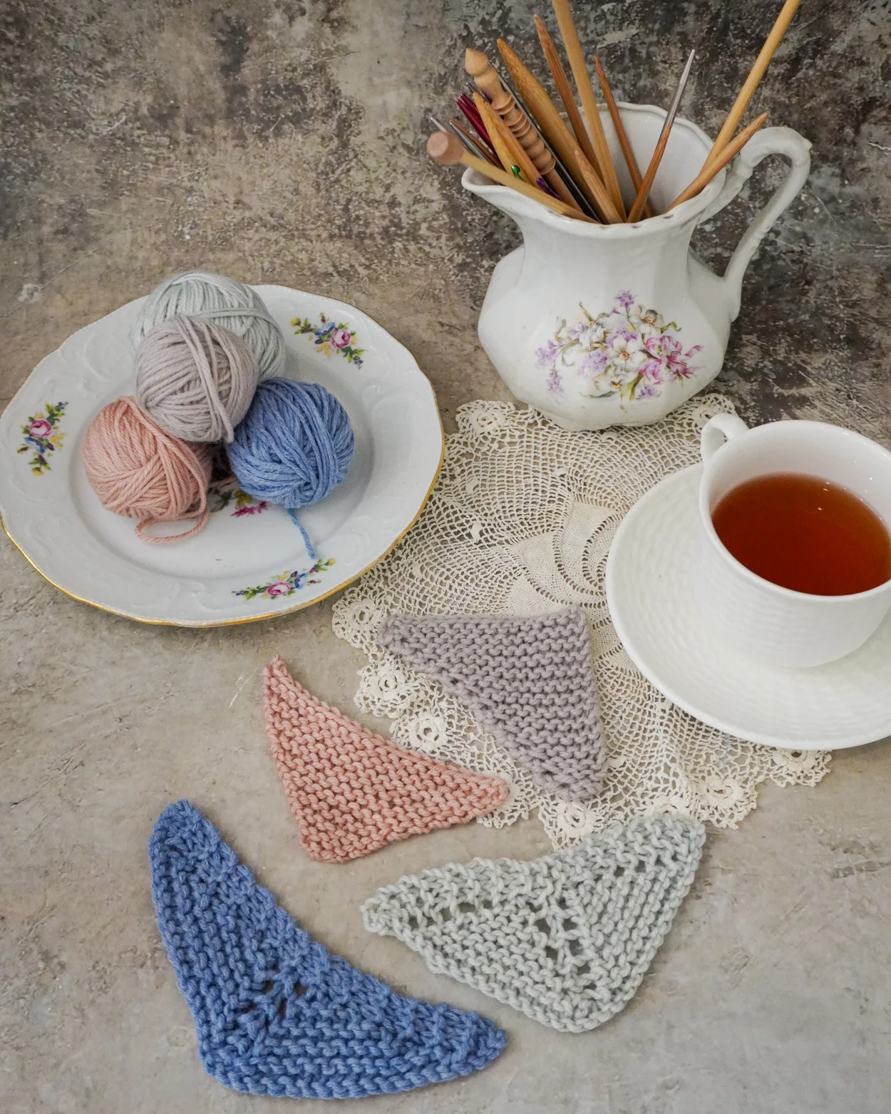 A flatlay photo showing four mini triangle shawls in slightly different shapes at the bottom center. They're surrounded by a plate full of mini yarn balls, an antique pitcher filled with knitting needles and crochet hooks, and a white teacup and saucer full of tea.