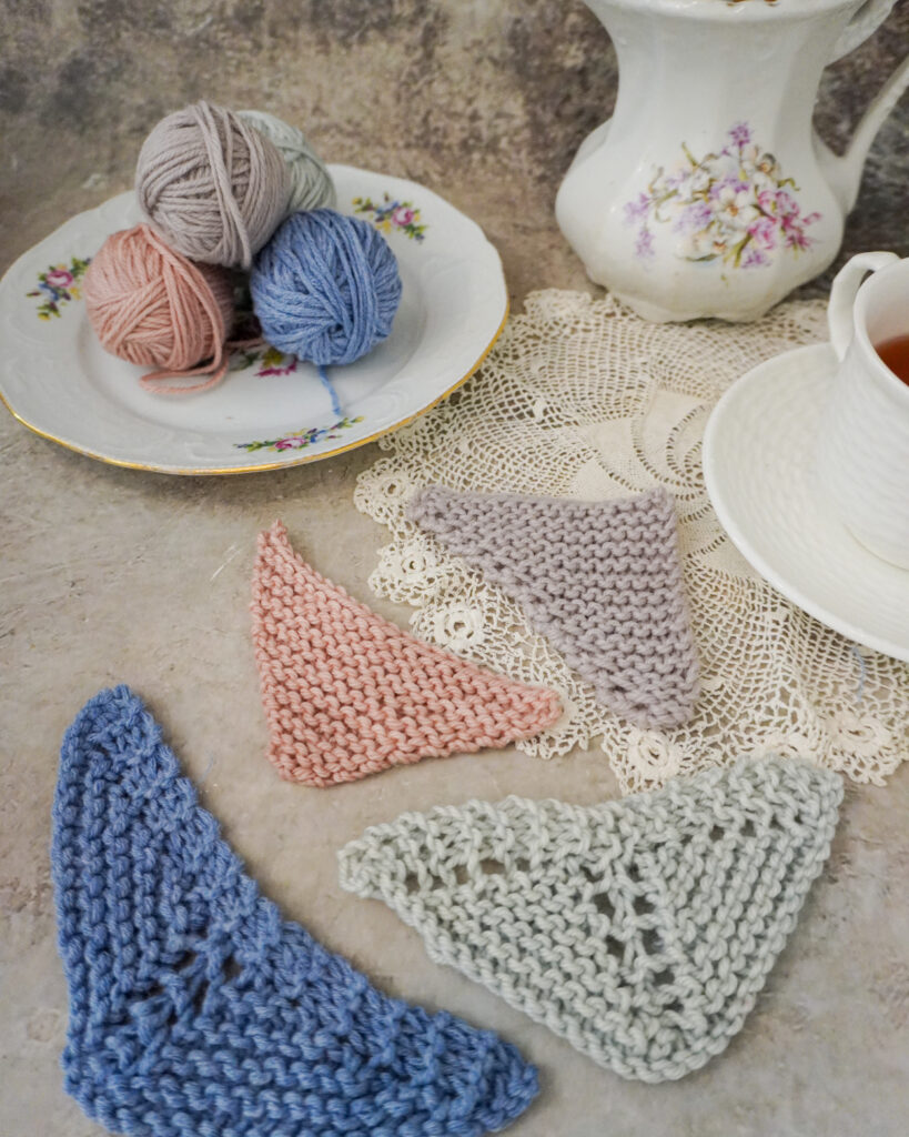 Four mini triangle shawls in different pastel shades of yarn fill the foreground of the image. In the background are a plate full of mini yarn balls, an antique pitcher, and a white teacup full of tea.