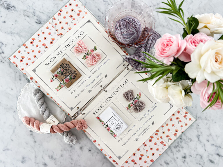 A horizontal image showing an open binder with two pages labeled "Sock Mending Log." Each page features an affixed yarn label and enough yarn for one or two mends of socks in the future. The binder is surrounded by a few mini skeins of yarn, a knit sock in progress, and a vase of pink and white flowers.