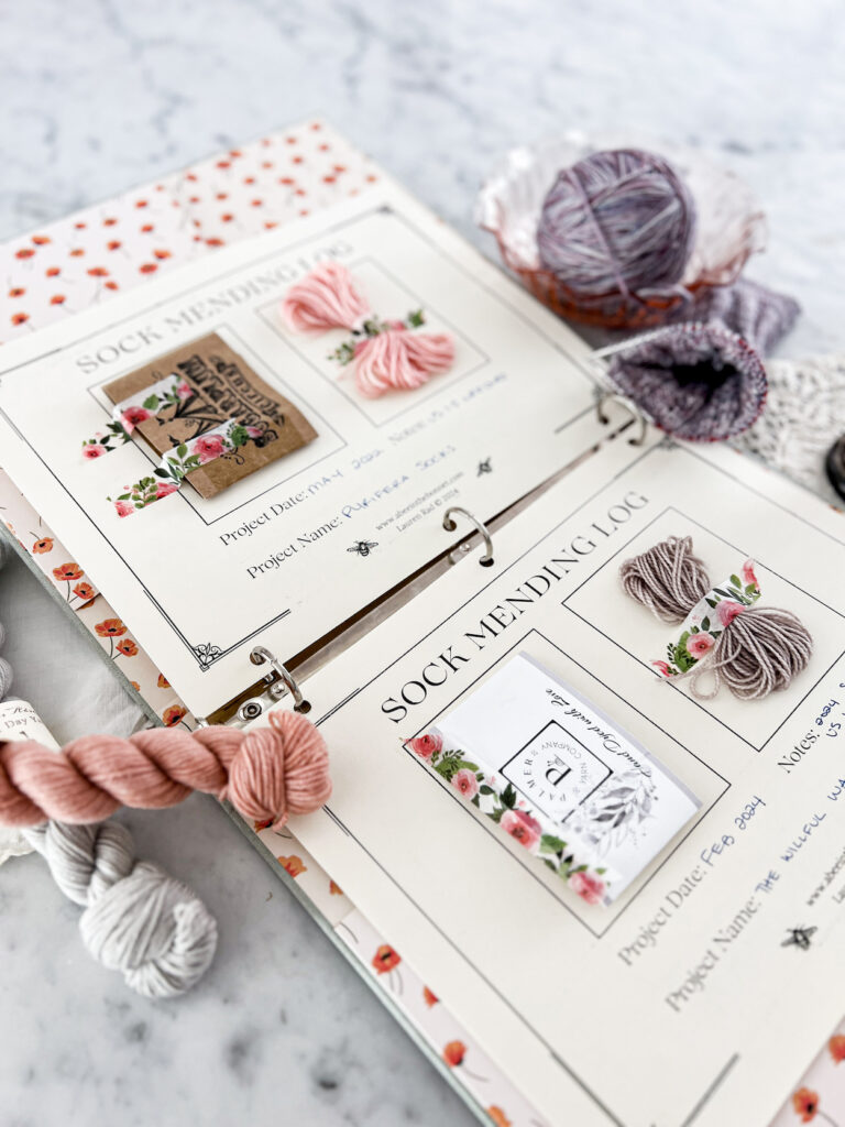 A low angle photo showing a close-up on a yarn label affixed to one page of a Sock Mending Log. The page is clipped into a binder and surrounded by mini skeins of yarn and a knit sock in progress.