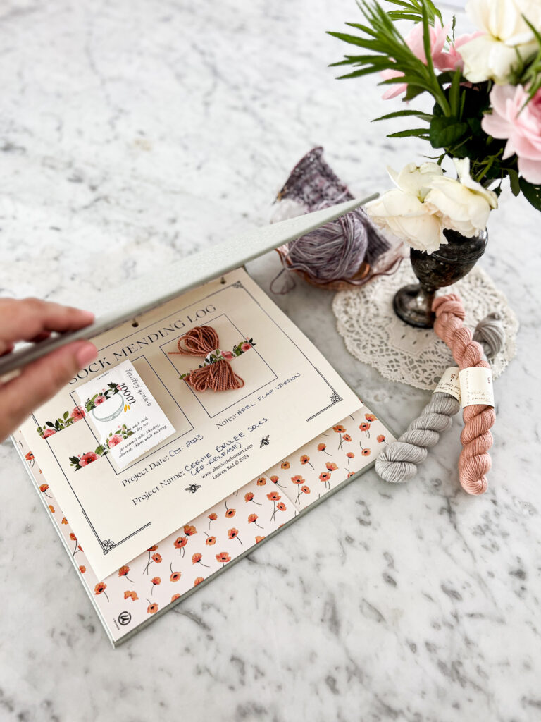 A white woman's left hand lifts the cover of a pale green binder to reveal a page inside. The page says "Sock Mending Log" across the top. A yarn label and small amount of yarn for future mending have been attached to the page.