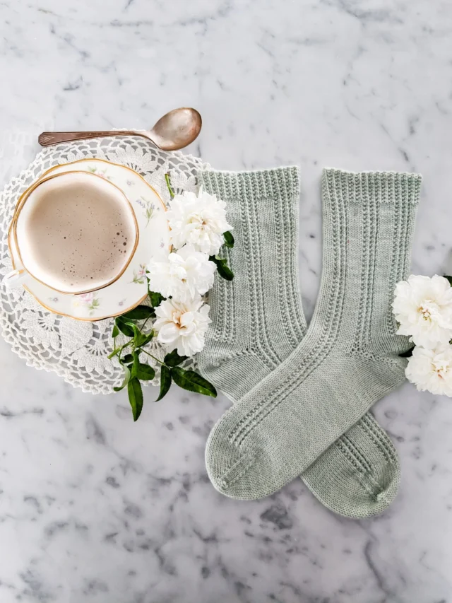 A top-down photo of a pair of light green handknit socks laid out on a white marble countertop. The socks have simple columns of purl stitches for textural decoration. Next to them are some white roses and a white teacup and saucer full of milky coffee.