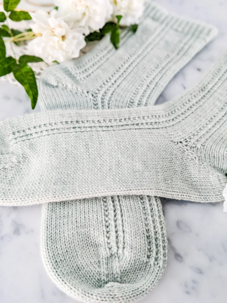 A pair of light green handknit socks are laid out on a white marble countertop. The socks are crossed at the feet. This photo focuses on the toes and feet of the socks.