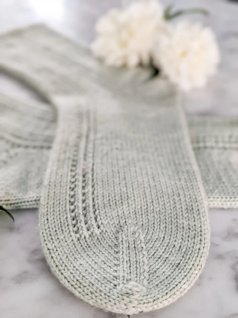 A zoomed-in photo focused on the toe of a handknit sock knit in a soft green color. The rest of the sock blurs away into the background.