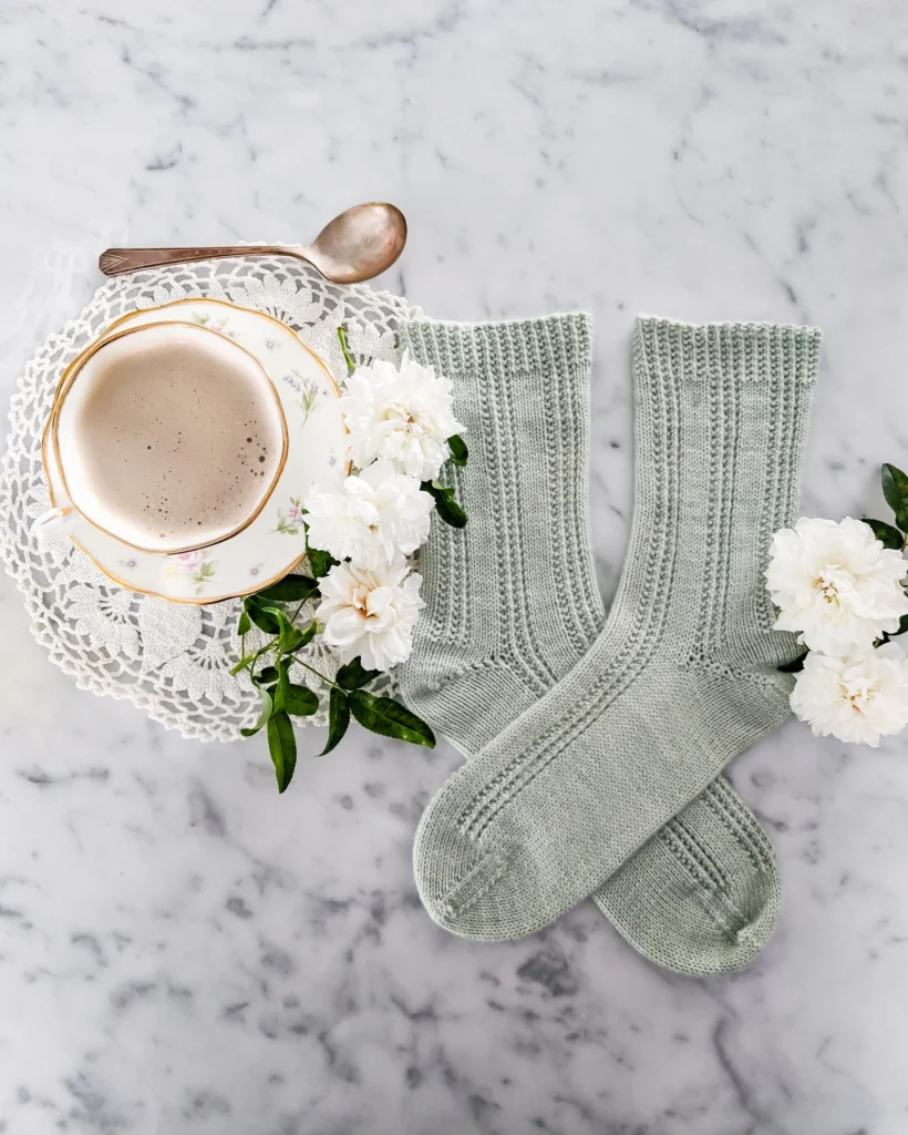 A top-down photo of a pair of light green handknit socks laid out on a white marble countertop. The socks have simple columns of purl stitches for textural decoration. Next to them are some white roses and a white teacup and saucer full of milky coffee.