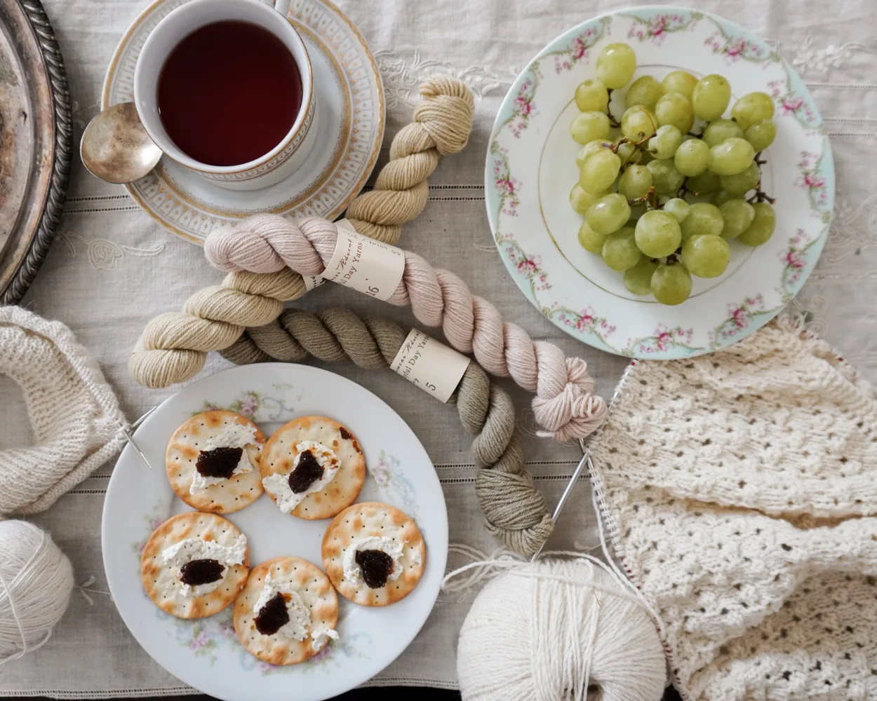 A horizontal image showing a plate of green grapes, a plate of crackers with cheese and fruit spread, and a teacup full of hot tea sit on a table surrounded by white knitting and mini skeins of pastel yarn.