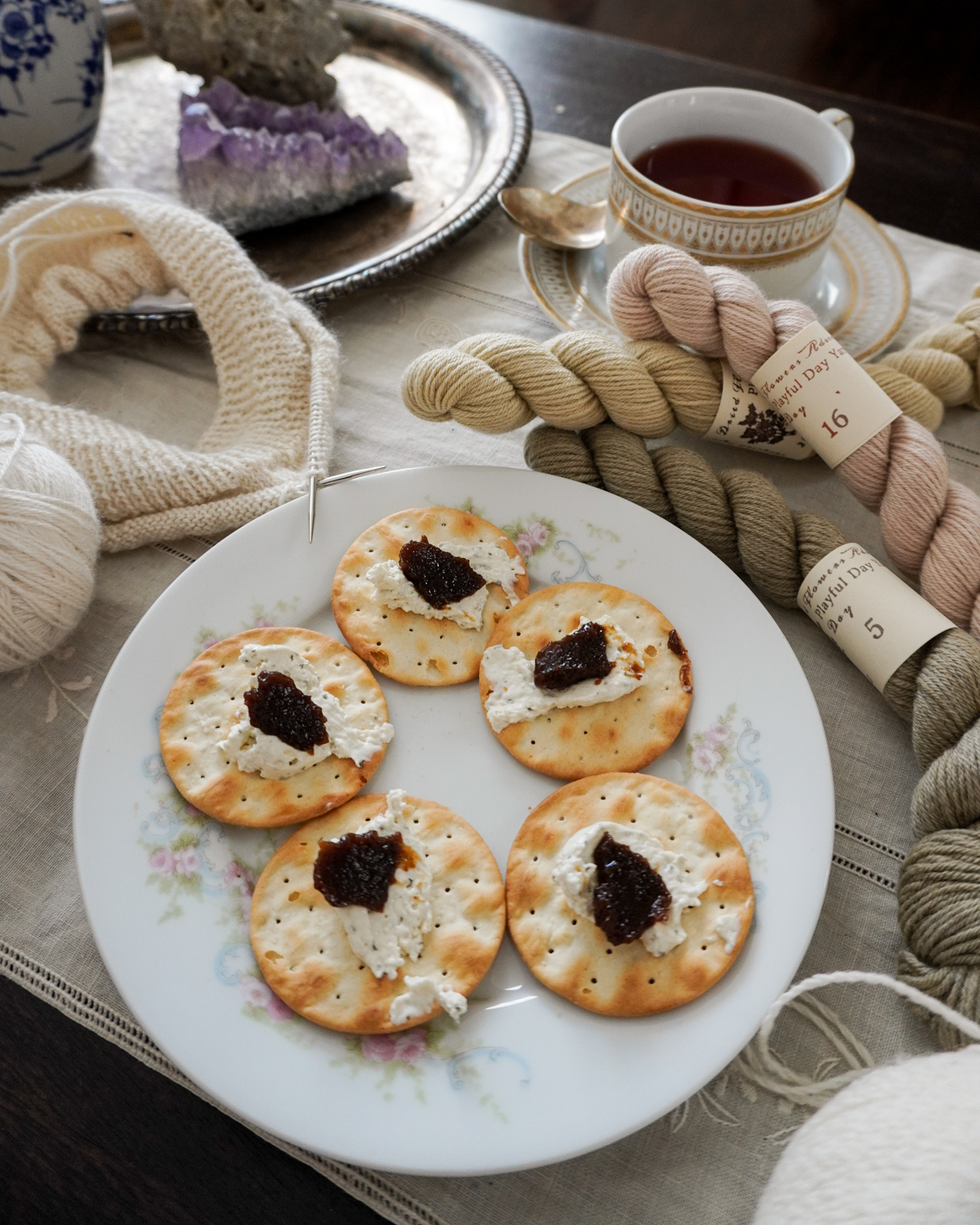 A pastel plate with five round crackers. Each cracker has some soft cheese and fruit spread on it. The plate is surrounded by white knitting and pastel skeins of yarn.