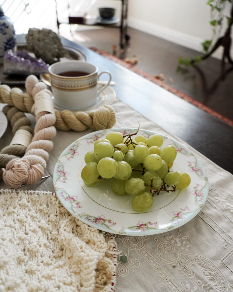 A plate of green grapes fills the lower right of the photo. Blurred in the background are mini skeins of yarn and tea.