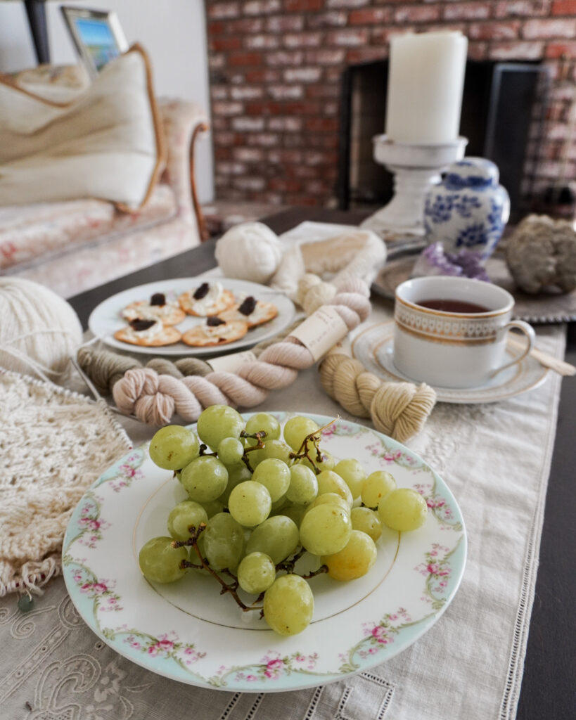 A pastel plate of grapes occupies the foreground of the image. Blurred in the background are more snacks, yarn, and tea.