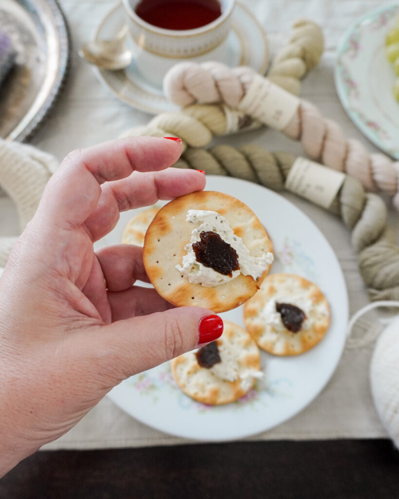 A white woman's left hand holds a cracker with some cheese and fruit spread on it.