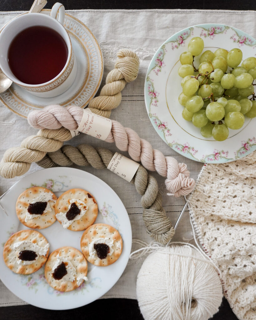 A plate of green grapes, a plate of crackers with cheese and fruit spread, and a teacup full of hot tea sit on a table surrounded by white knitting and mini skeins of pastel yarn.