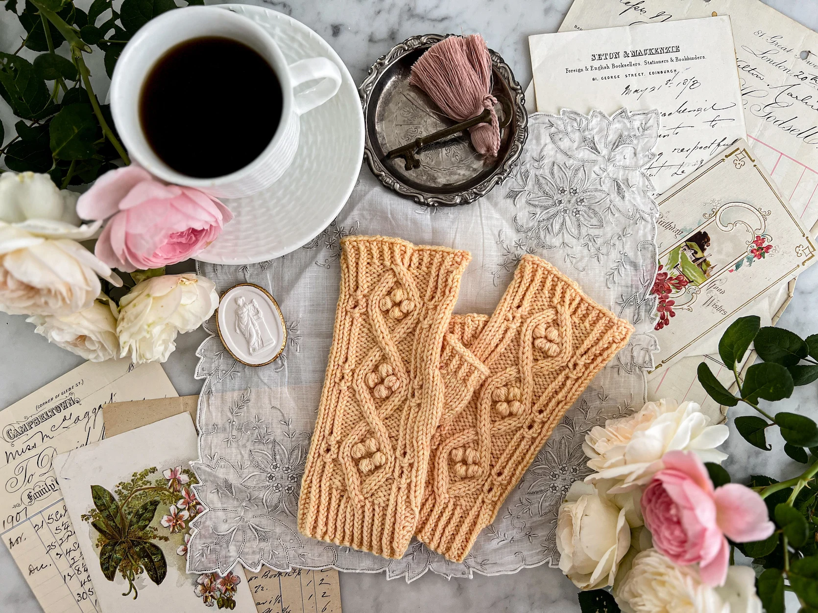 A pair of yellow fingerless mitts is laid out flat on an antique handkerchief surrounded by a teacup full of coffee, white and pink roses, and antique paper ephemera. The mitts are full of texture and have a thumb gusset for a comfortable fit.