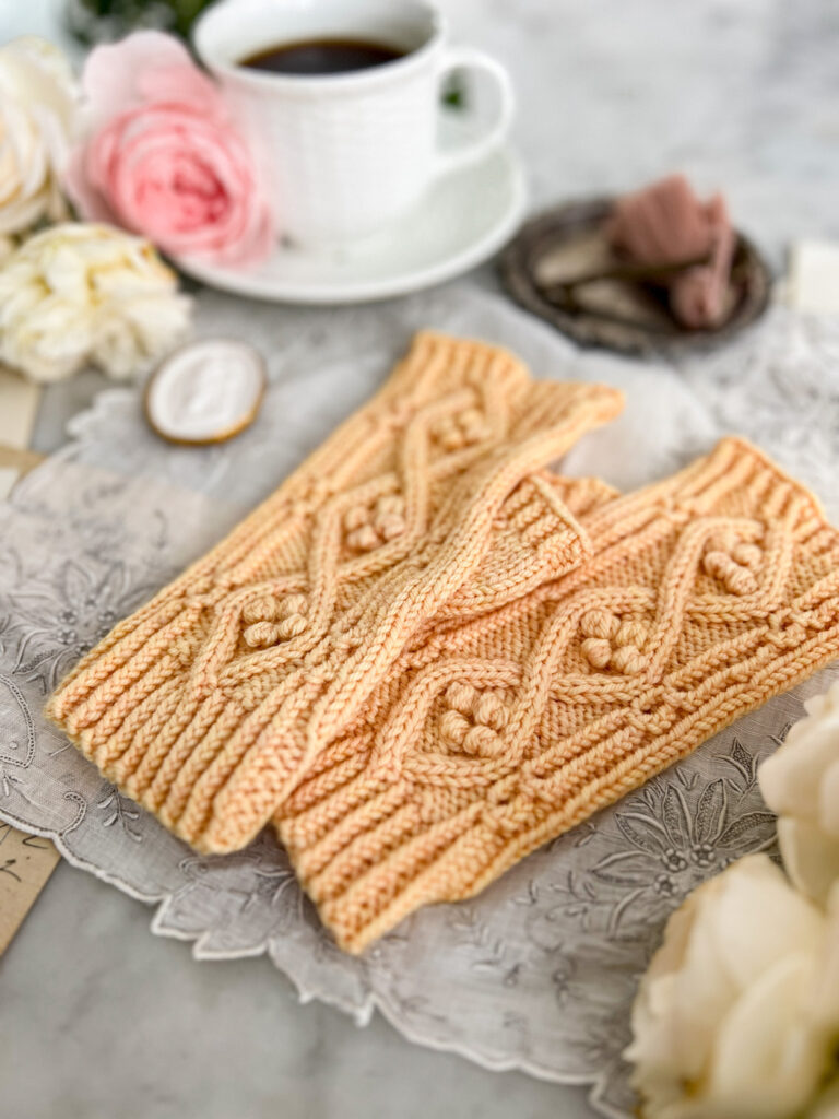 A pair of yellow handknit fingerless mitts sits on an antique handkerchief. The mitts are photographed from a low angle focused on the bottom cuffs. The mitts fade away out of focus as the image moves toward the fingertips. The intricate texture on the mitts features coin lace, twisted ribbing, cables, and bobbles nestled inside the cables.