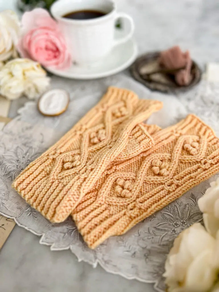 A pair of yellow handknit fingerless mitts sits on an antique handkerchief. The mitts are photographed from a low angle focused on the bottom cuffs. The mitts fade away out of focus as the image moves toward the fingertips. The intricate texture on the mitts features coin lace, twisted ribbing, cables, and bobbles nestled inside the cables.