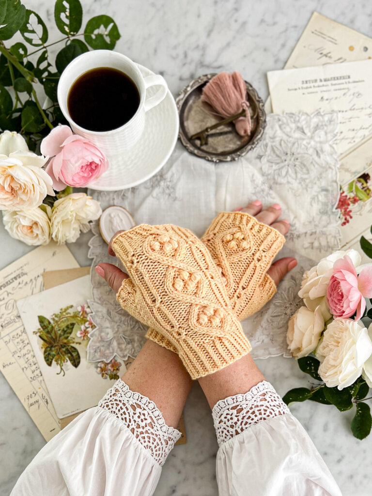 A white woman's hands are crossed at the wrists, right over left, on a white marble countertop surrounded by antique paper ephemera. She is wearing a pair of yellow handknit fingerless mitts that are full of texture, including cables, coin lace, twisted ribbing, and bobbles nestled inside the cables.