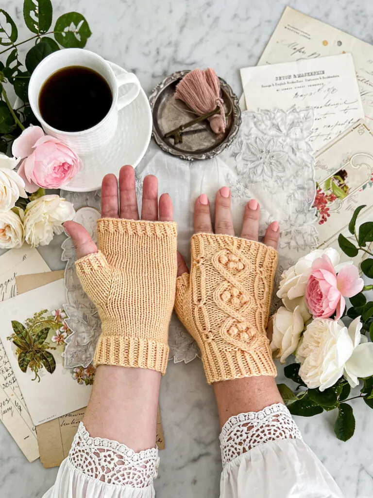 A white woman's hands are resting on a white marble countertop, one hand palm up and one hand palm down. This photo shows the top and bottom of the Daffodils in Sunshine Mitts, a pair of yellow handknit mitts featuring lots of texture.