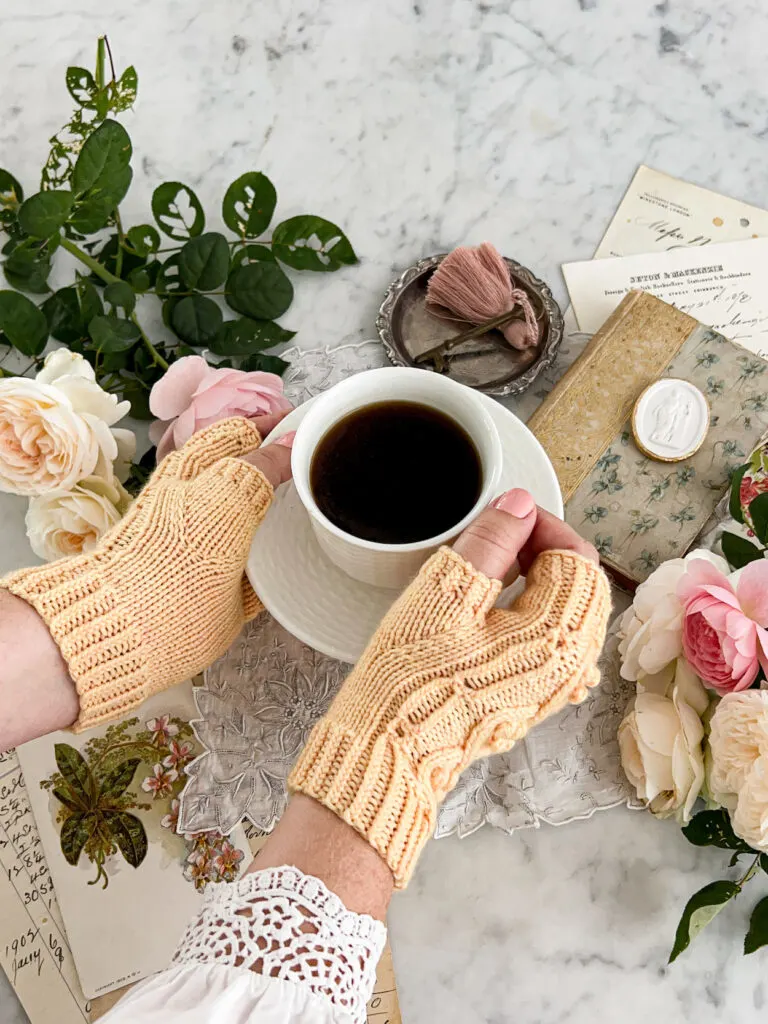 A white woman's hands wearing a pair of yellow handknit fingerless mitts lift a white teacup and saucer. The teacup is filled with black coffee. In the background are lots of roses and antique paper ephemera.