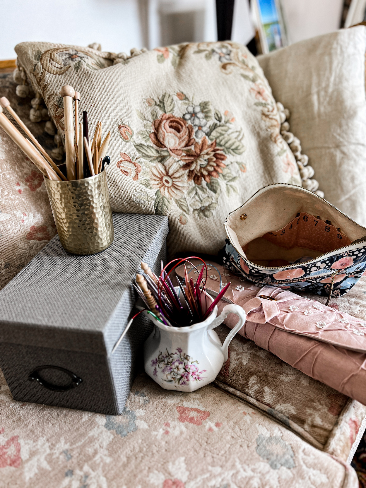 A collection of tools for organizing knitting needles, including a special pouch with organizer slots, a pink needle roll, an antique white pitcher, a lidded box, and a hammered brass cup are displayed together on an old sofa with pastel floral upholstery.