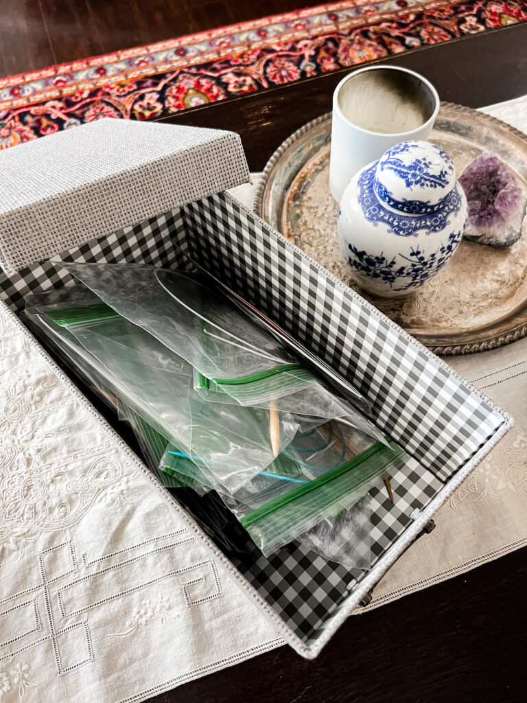 A gray lidded box sits on a tabletop with the lid mostly off the box, showing lots of zippered plastic baggies with knitting needles in them inside the box.