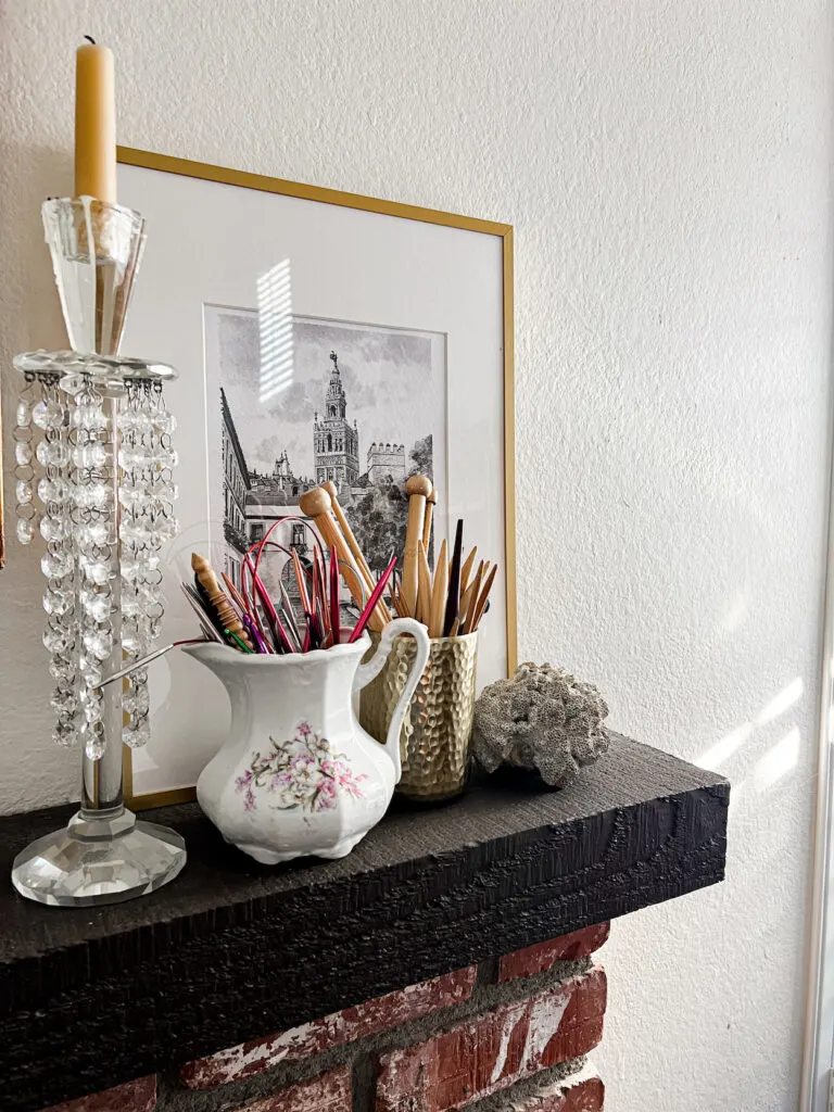 An antique stoneware pitcher and hammered brass cup, both full of knitting needles, sit on a dark wooden mantel surrounded by coral, a framed print, and a crystal candlestick.