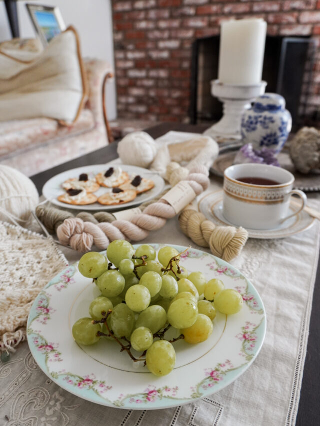 A pastel plate of grapes occupies the foreground of the image. Blurred in the background are more snacks, yarn, and tea.