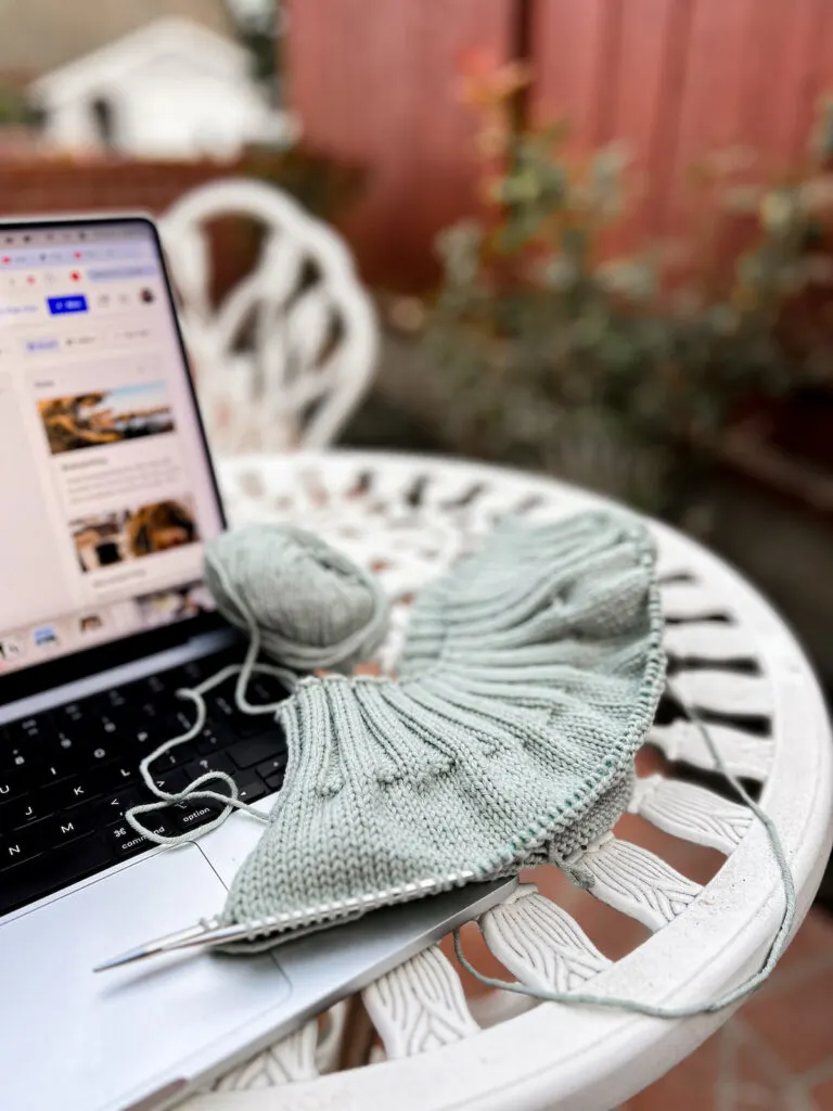 A light green knit sweater in progress sits outdoors on a white cast iron table next to an open laptop.