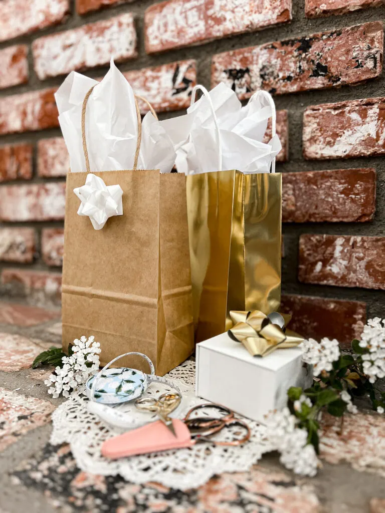 A pile of gifts sit on a brick fireplace hearth. There are two gift bags with tissue paper peeking out the top, a white box with a gold bow on it, and some scissors and measuring tape.