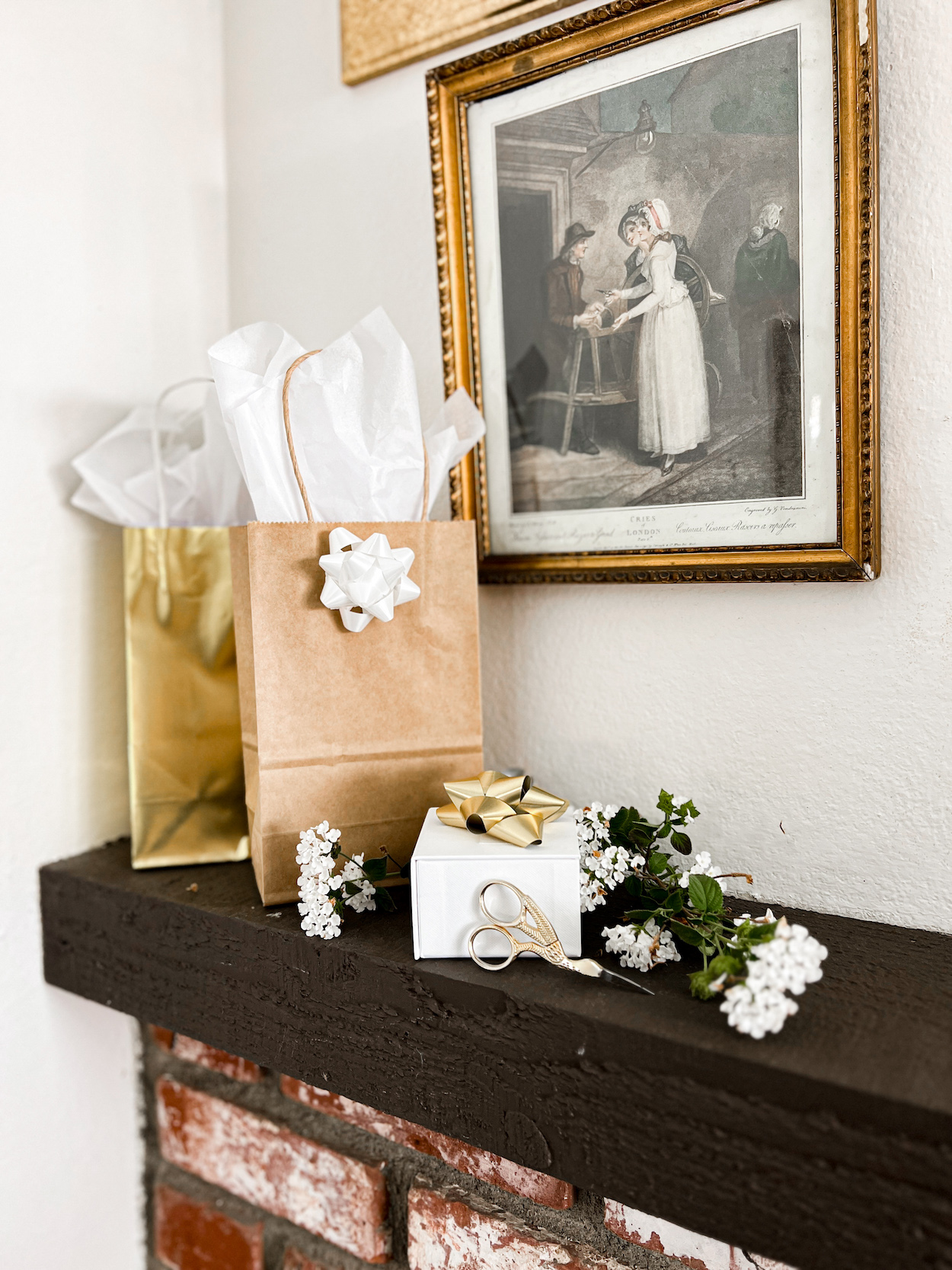 A couple gift bags, a white gift box with a bow on top, some white lantana, and a pair of gold scissors sit on a dark wood mantel above a brick fireplace.