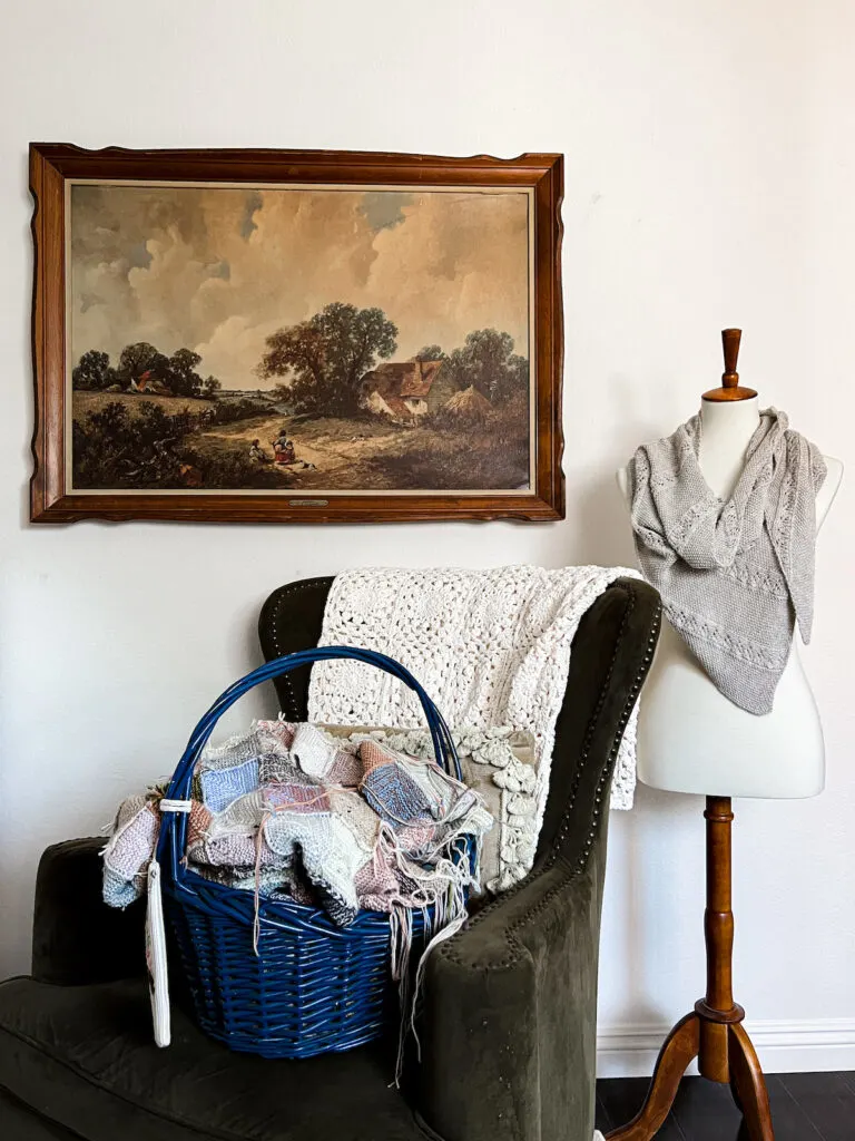 A blue basket full of knitting sits on a green wingback armchair. In the background are a large landscape print and a white dressmaker's form with a shawl on it.