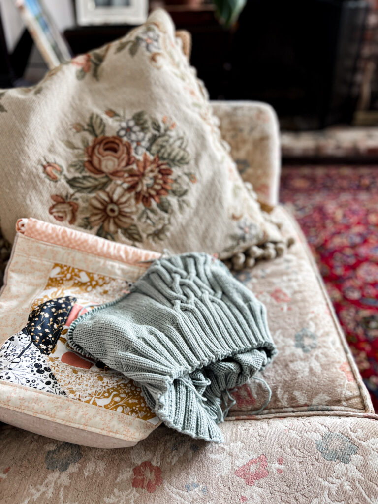 A pale green knit sweater in progress sits on a vintage sofa with a project bag next to it.