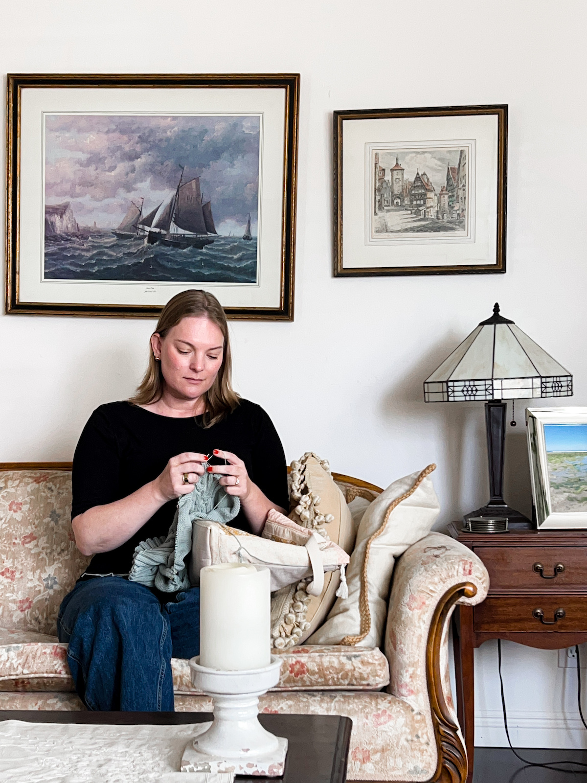 A white woman with shoulder-length hair sits on a vintage sofa. She is wearing a black tshirt and jeans, and she's knitting a pale green sweater.