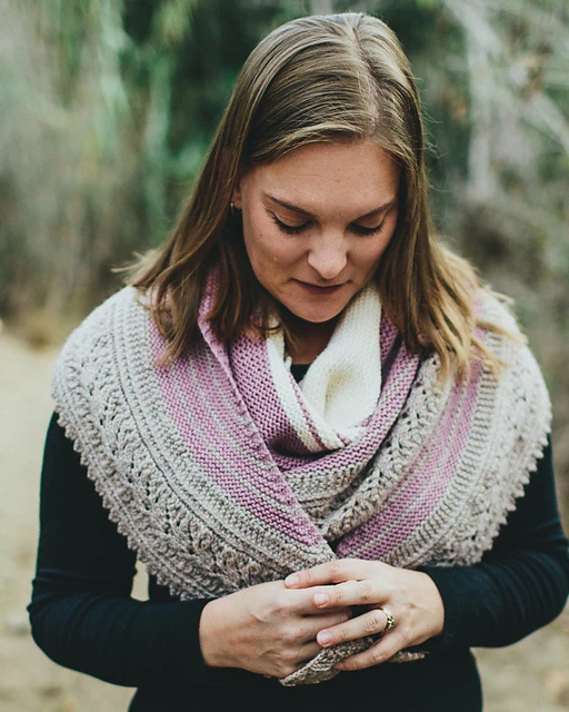 A white woman with dark blonde hair looks down at her hands, which are folded across the ends of a crescent-shaped shawl that is wrapped multiple times around her shoulders. The shawl is a crescent-shaped shawl in cream, lavender, and light brown, and the light brown part is an intricate lace border.