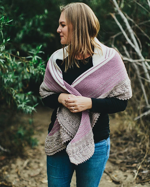 A white woman with dark blonde hair wears blue jeans and a black, long-sleeved tee-shirt with a handknit shawl wrapped around her shoulders. She's looking off to the side and is surrounded by shrubs and trees. The shawl is a crescent-shaped shawl in cream, lavender, and light brown, and the light brown part is an intricate lace border.