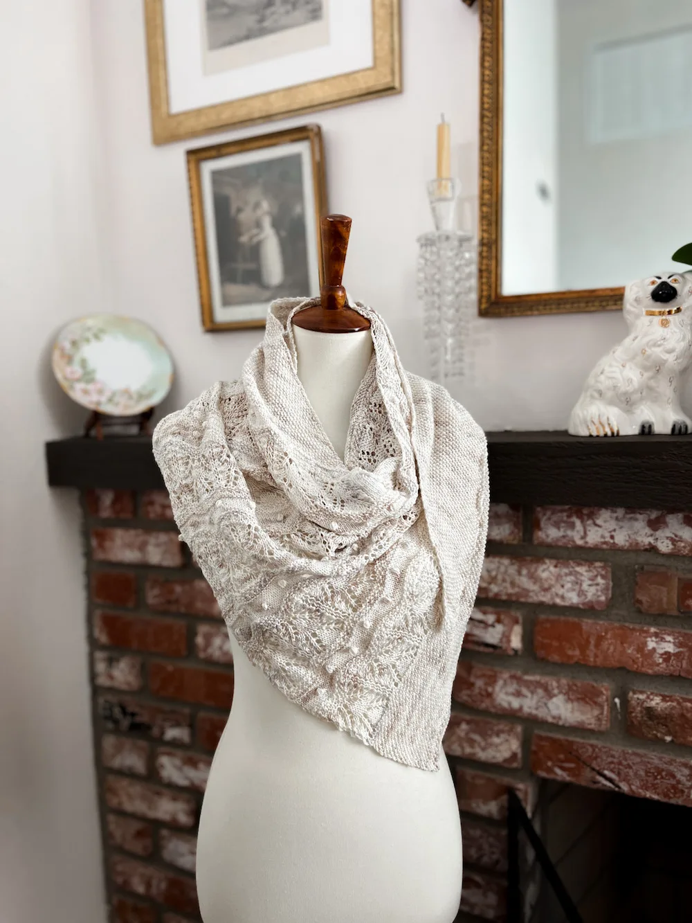 A white shawl with gray and tan variegations is draped around the shoulders of a dressmaker's form. This is a head-on photograph that shows how the shawl would look when worn with the point draped toward the front of the body. You can see the seed stitch bands, lace, and tiny bobbles that make up the different textures of the shawl.