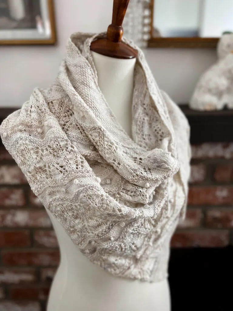 A side view of a white shawl with tan and gray variegations draped around a dressmaker's form. From this angle, you can more clearly see the tiny little bobbles and stripes of lace that make up the textured portion of the shawl.