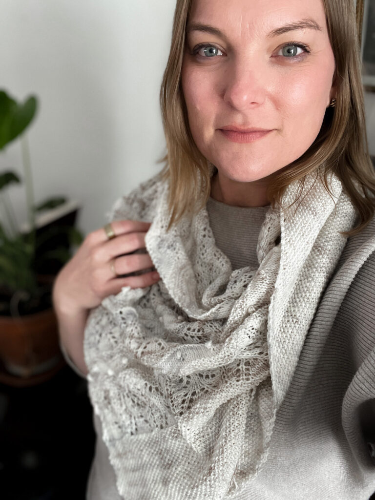 A white woman in her late 30s (me!) smiles softly at the camera while wearing a handknit shawl in shades of cream, gray, and tan. The shawl is draped so that the point hangs down in front.