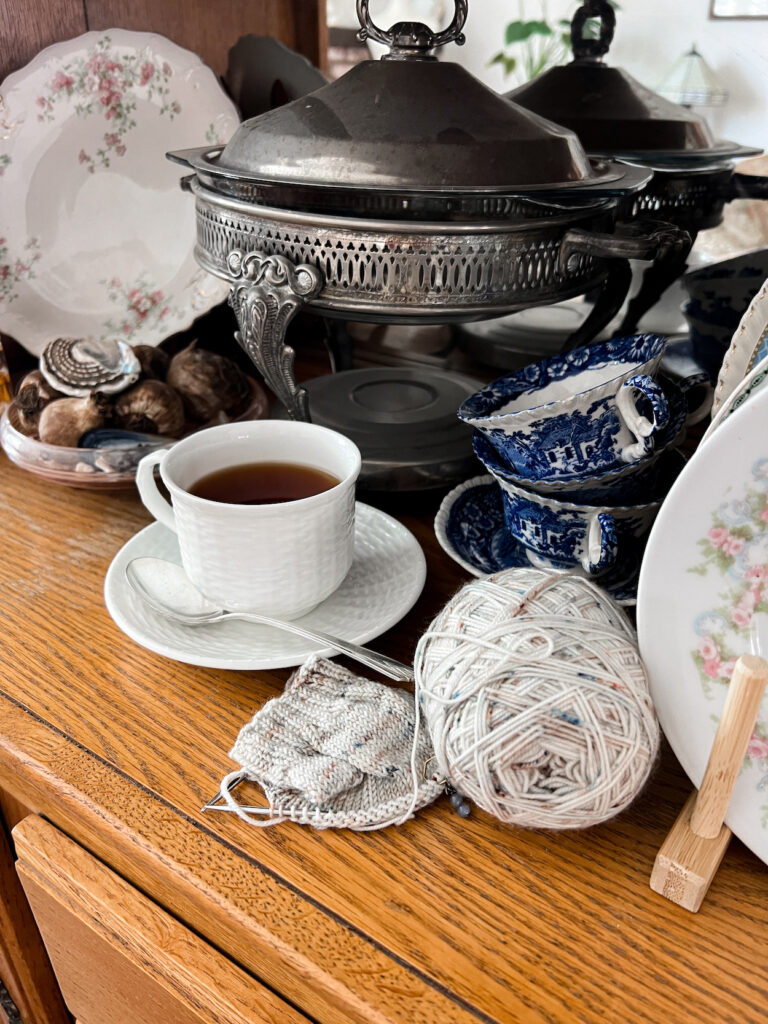 A handknit sock in progress sits on a shelf of a china hutch full of vintage dishes, seashells, and bric a brac.