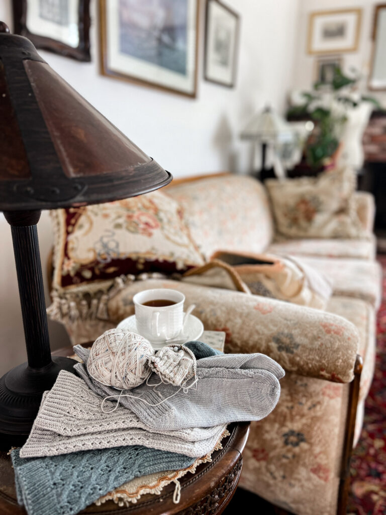 A pile of handknit gray and blue socks sits on a side table. In the background is a faded vintage sofa with needlepoint pillows and a wall full of vintage prints.