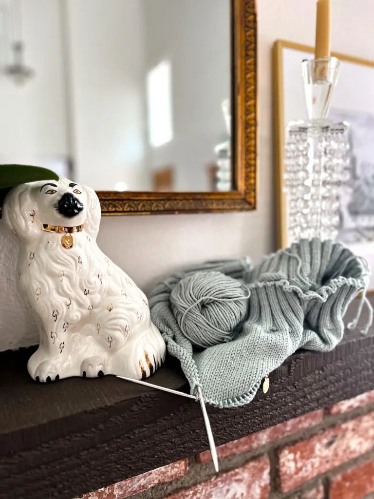 A soft green handknit sweater on the needles perches on a dark brown mantel between a white Staffordshire dog figurine with a judgmental facial expression and a crystal candlestick.