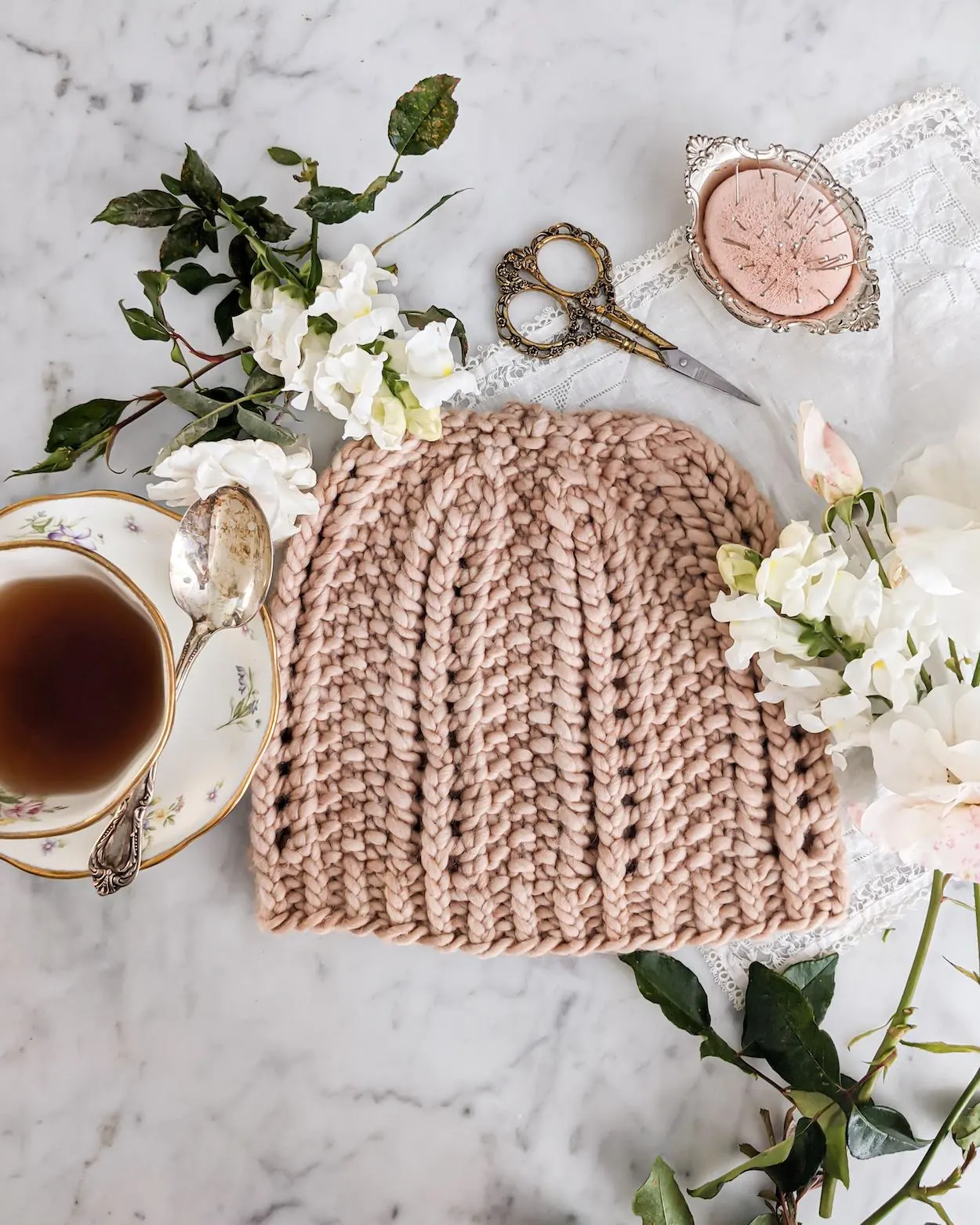 A flatlay photograph of the Pebbles by the Sea Hat. The hat, a chunky-weight handknit hat in a clay-colored yarn, is laid out on a white marble countertop and surrounded by antique bric a brac, white flowers, and a white teacup full of tea.