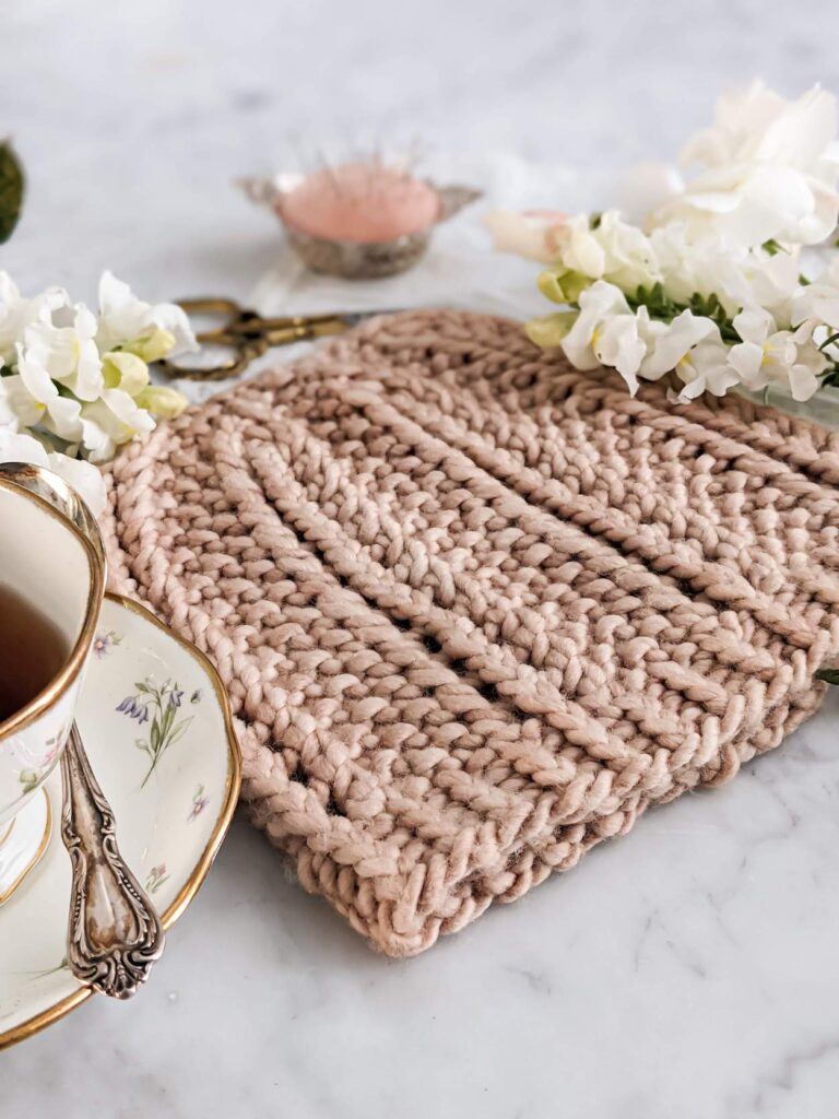 A close-up on the brim of the Pebbles by the Sea Hat. It shows the 1x1 twisted ribbing on the brim. The hat is knit in a clay-colored yarn and surrounded by white flowers on a white marble countertop.