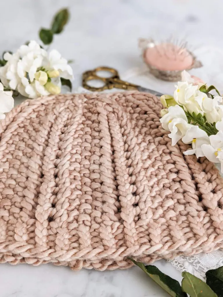 A close-up on the bottom of the Pebbles by the Sea Hat. It shows the 1x1 twisted ribbing on the brim, the seed stitch columns, and the columns of simple eyelets that add texture to the hat. The hat is knit in a clay-colored yarn and laid flat on a white marble countertop.