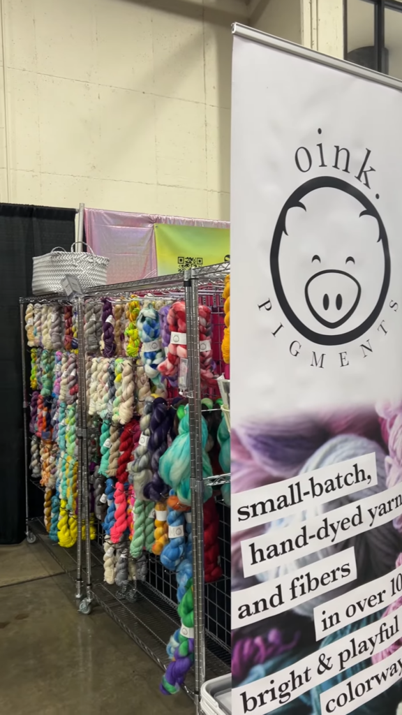 Part of the Oink Pigments booth at SoCal Fiber Fair 2024. There is a tall banner with their name and logo on the end of several wire shelves full of colorful twisted hanks of yarn.