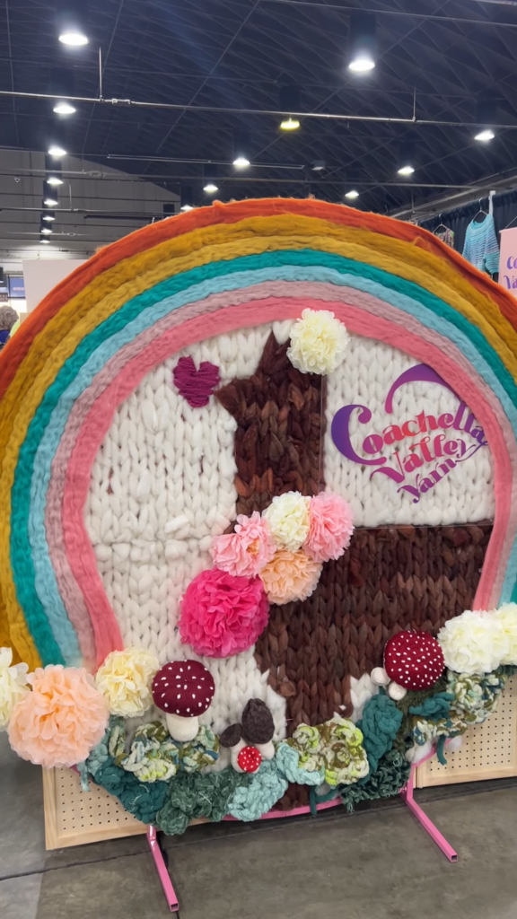 A giant fiber art display featuring a colorwork knit alpaca surrounded by a yarn rainbow. At the bottom are lots of faux flowers and mushrooms. The display says "Coachella Valley Yarn" in purple and pink letters.